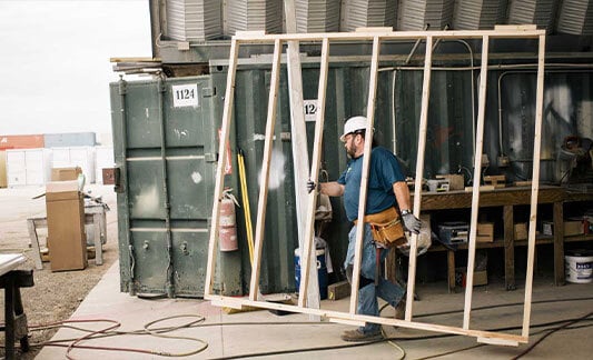 Worker moving a wood wall frame in front of shipping containers.